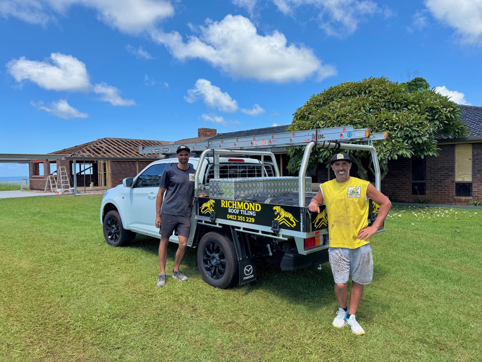 Two people in work clothes stand next to a utility truck parked on grass in front of a brick house under a blue sky.