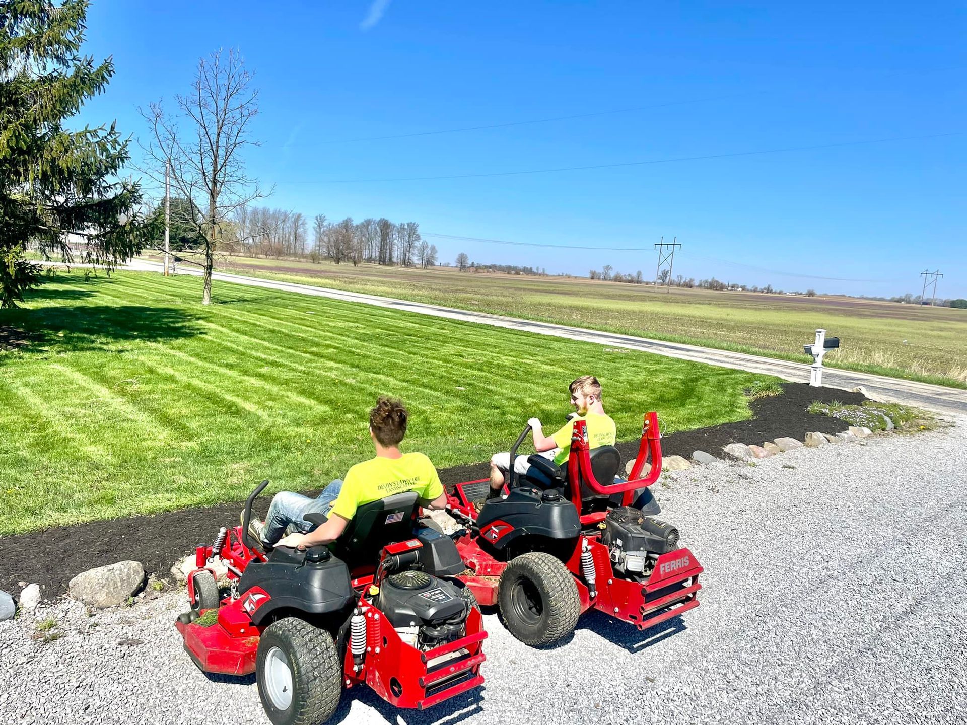 Two people are riding lawn mowers in a driveway.