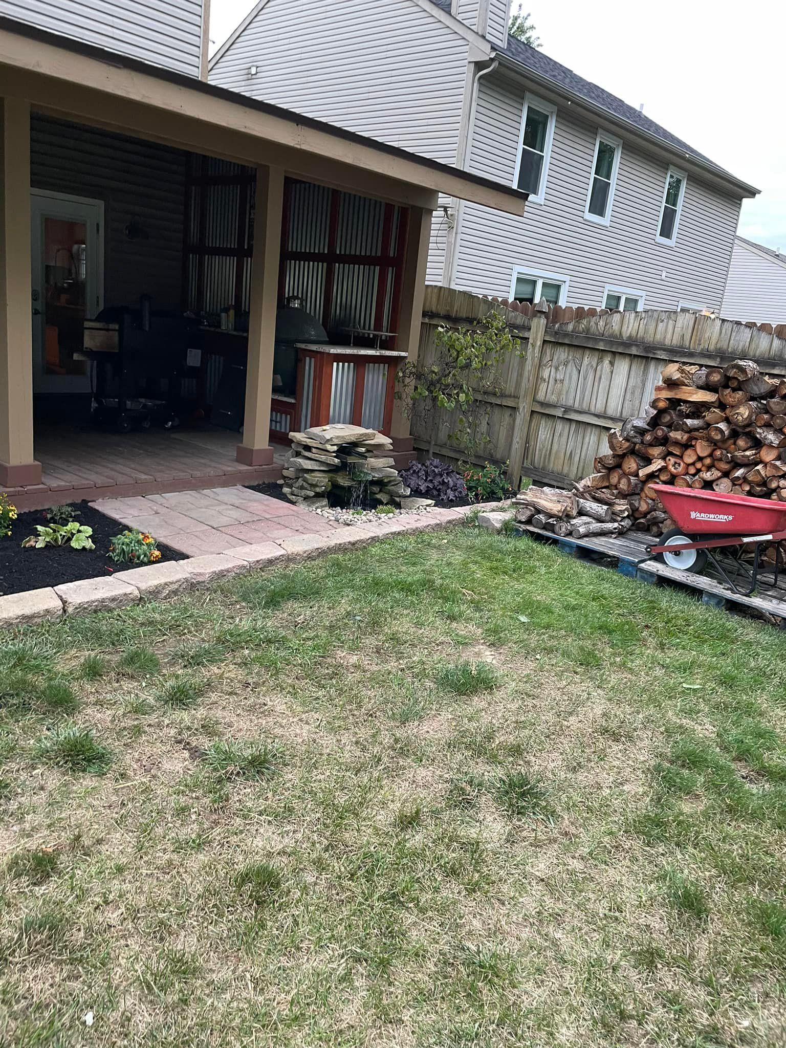 A backyard with a pile of wood and a wheelbarrow in front of a house.
