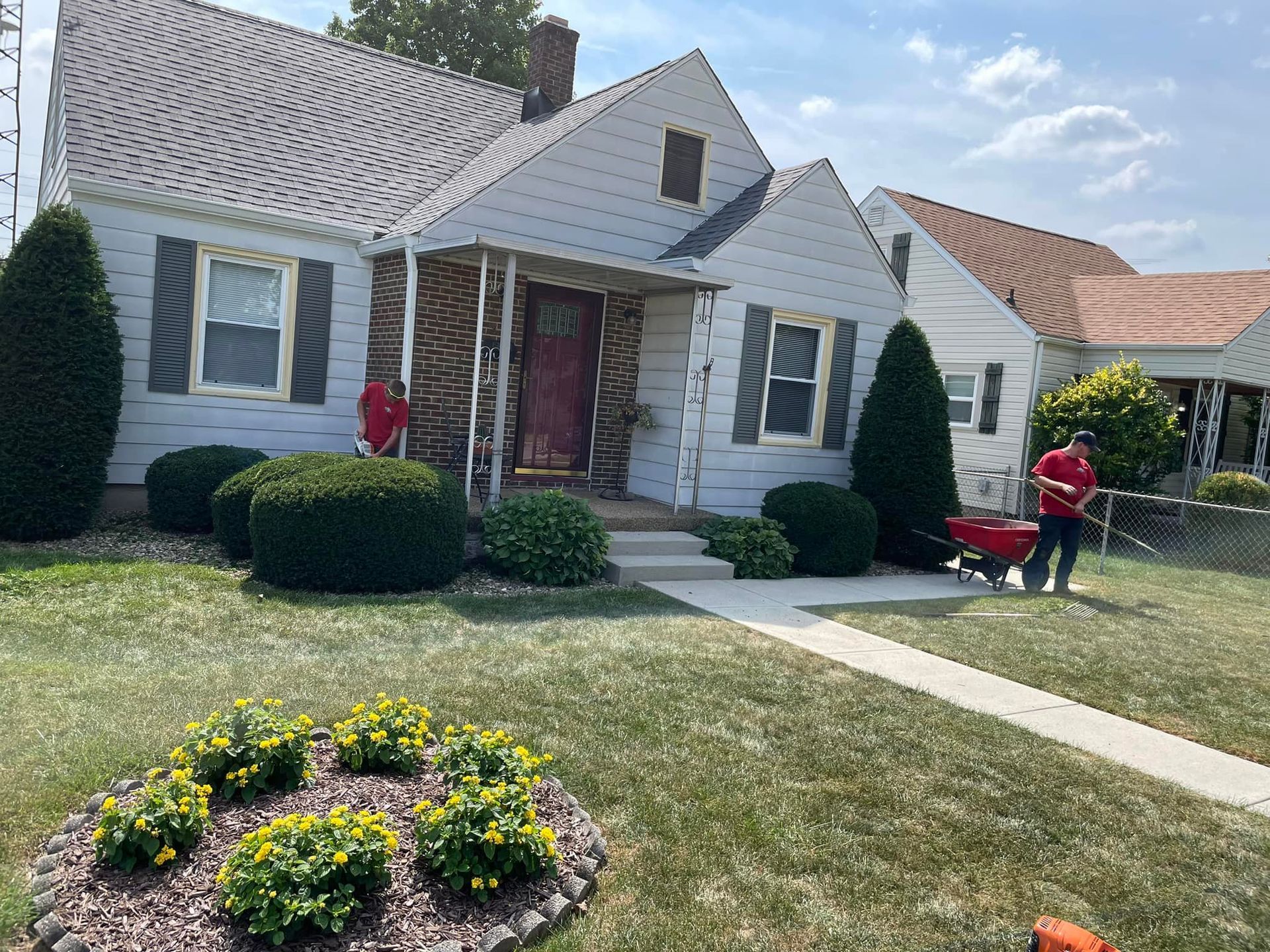 A man is standing in front of a white house with a red wheelbarrow.