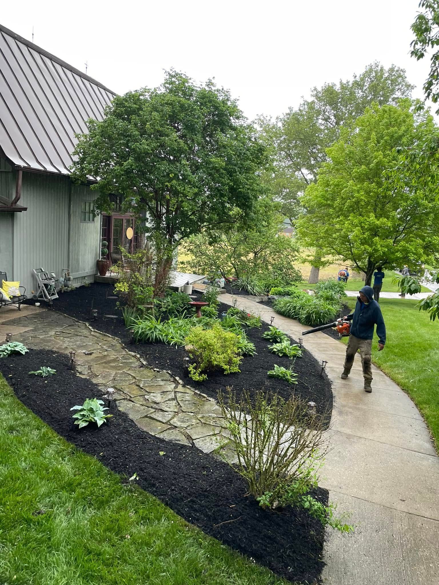 A man is blowing mulch in a garden in front of a house.