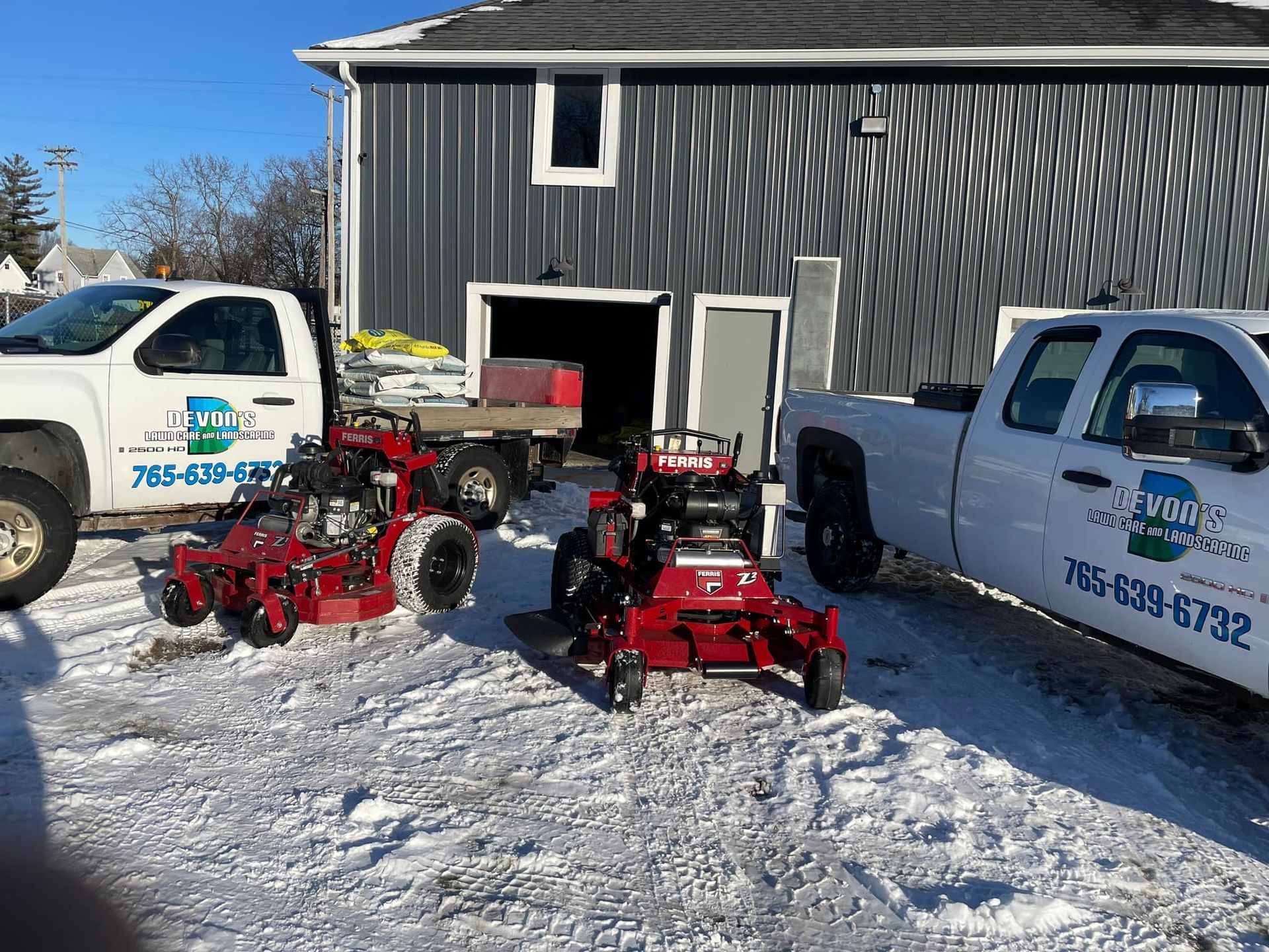 Two lawn mower trucks are parked in the snow in front of a building.