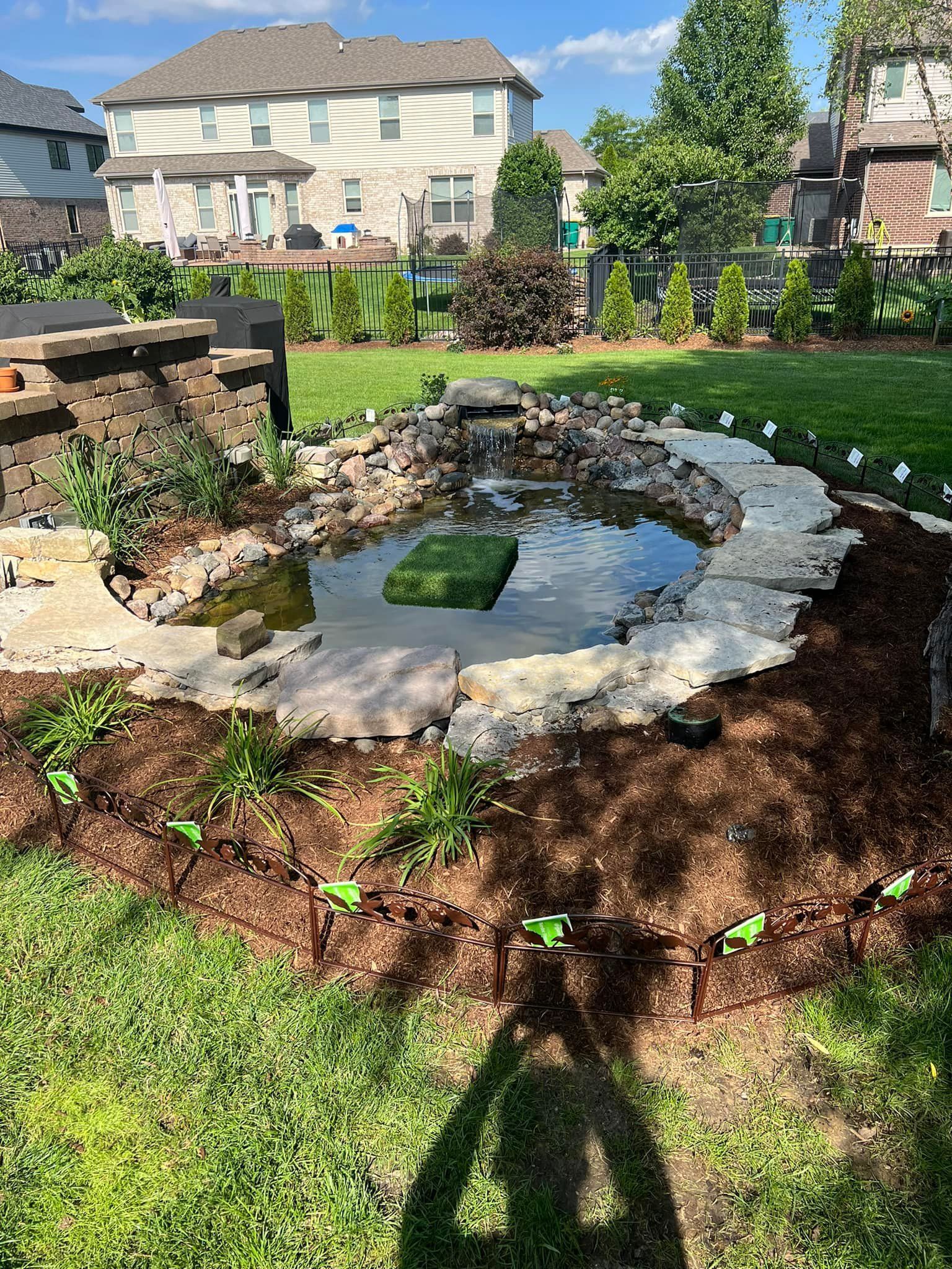 A backyard pond with a waterfall, surrounded by stones, plants, and mulch. A large house is
visible