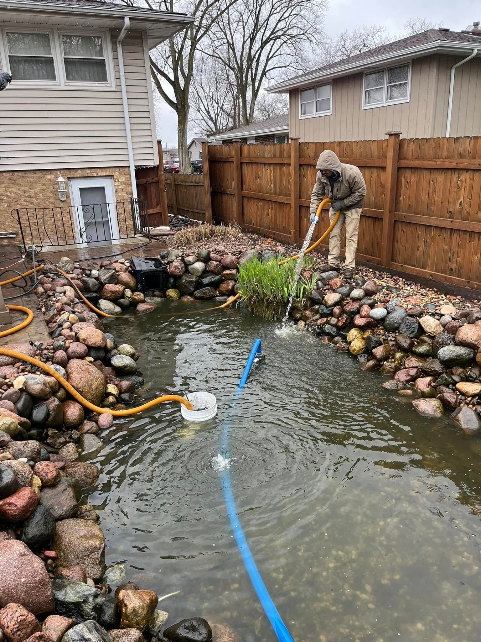 Person cleaning a backyard pond with a pump and hose, surrounded by rocks and a wooden fence.