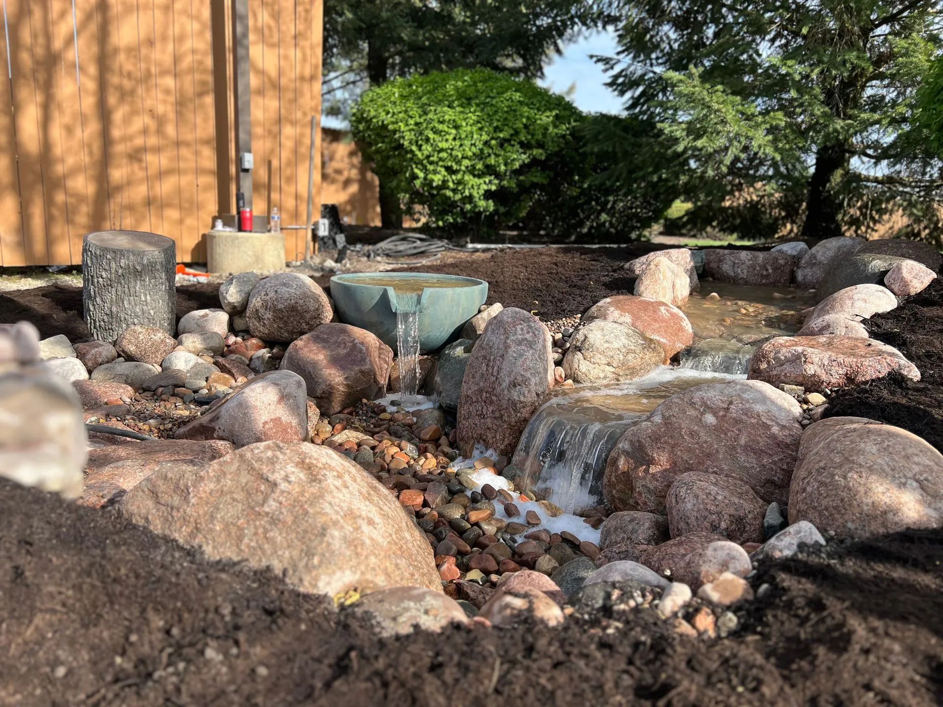 A small garden pond with a water feature, stone borders, and lush green bushes in a landscaped backyard.