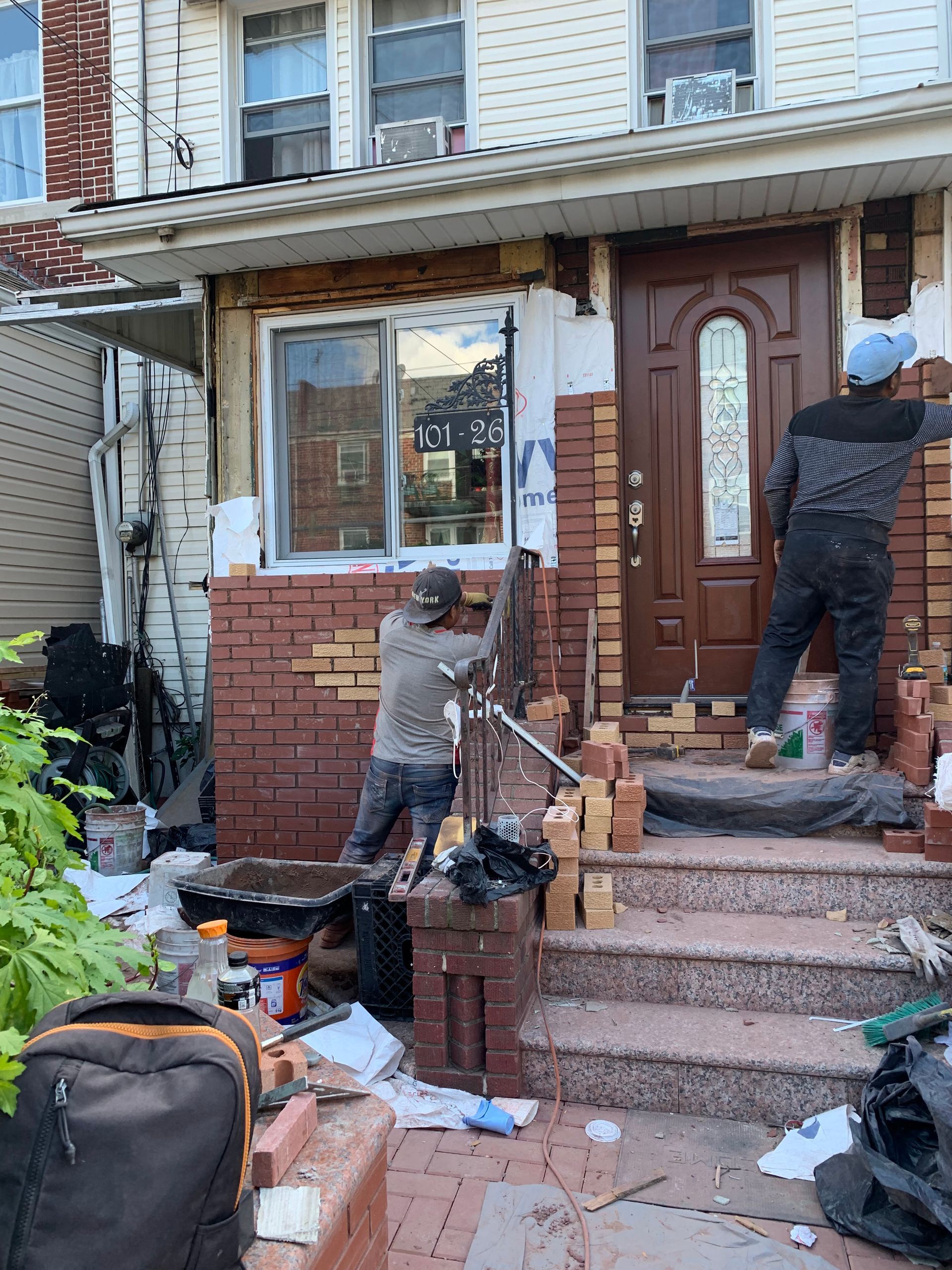 Two people working on a home exterior, installing brick facade and trim around a brown front door and window.