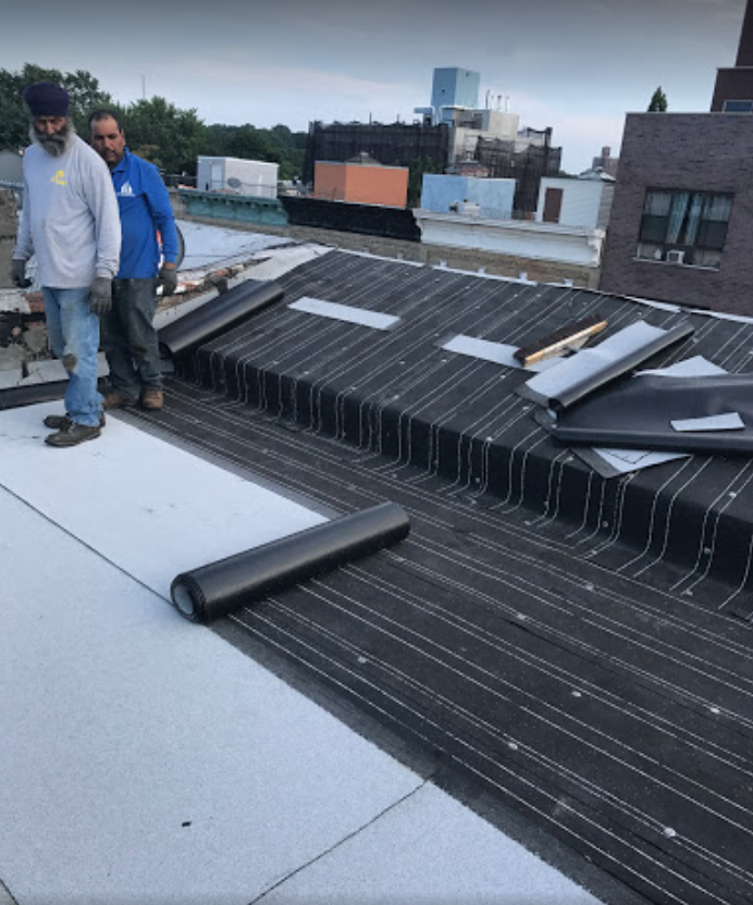Two men standing on top of a roof with rolls of roofing material on it