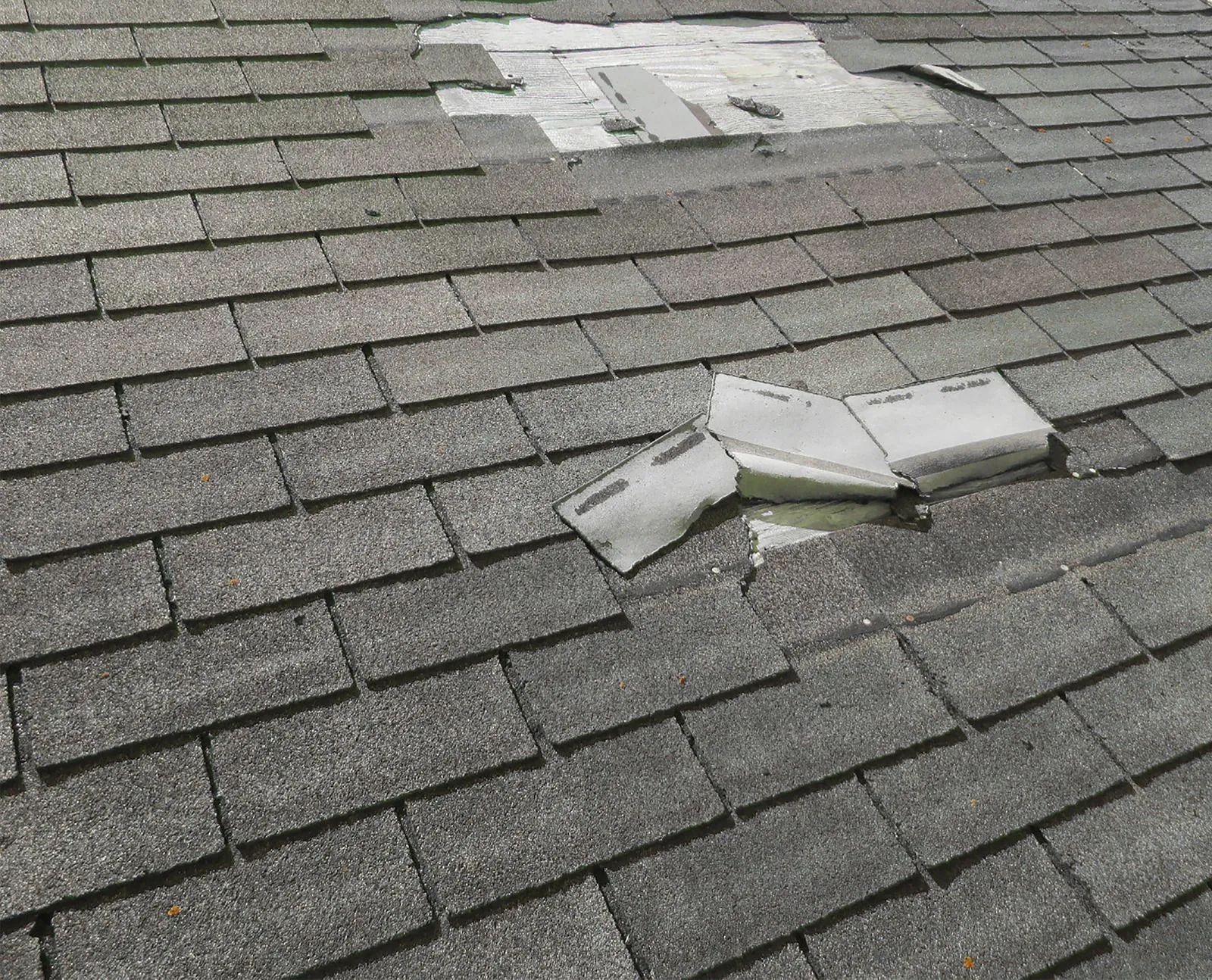 Damaged asphalt shingle roof with missing shingles. Gray, weathered texture with visible underlayment.