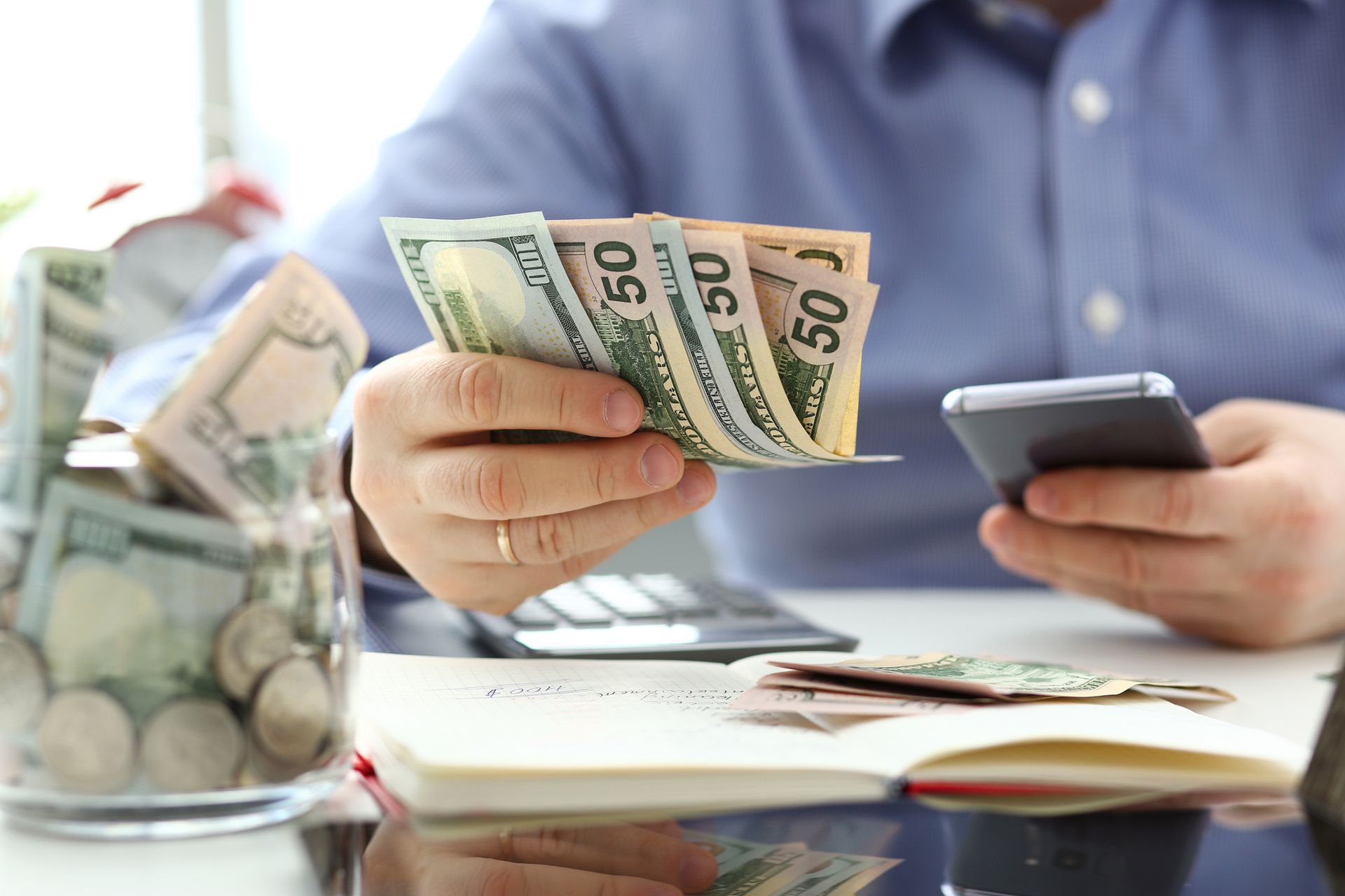 Man counting cash, with jar of money and smartphone on a desk.