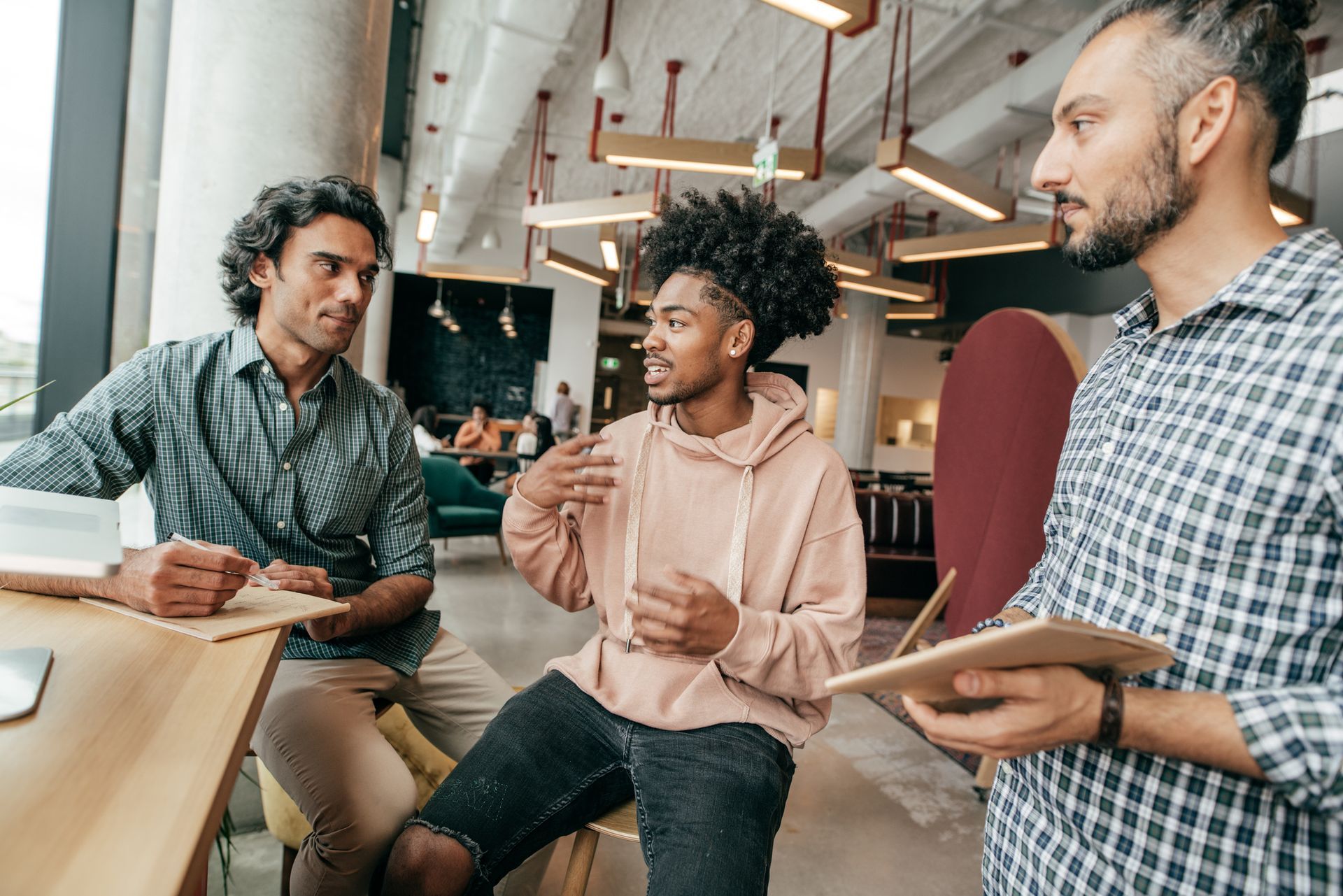 Three men in modern office setting, conversing. One man holds a tablet, gesturing.