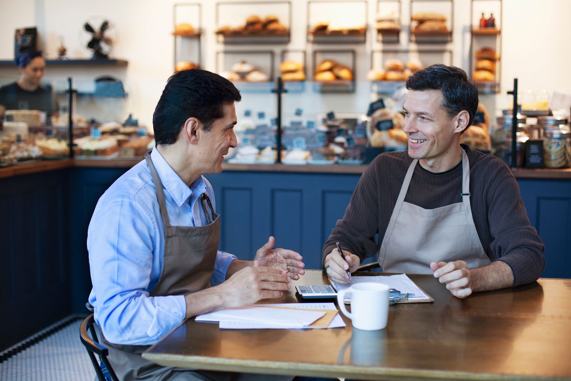 Two men in aprons, at a cafe table, talking and looking at papers, with a barista in the background.