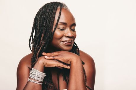 Smiling Black woman with graying dreadlocks, arms crossed on a table, eyes closed.