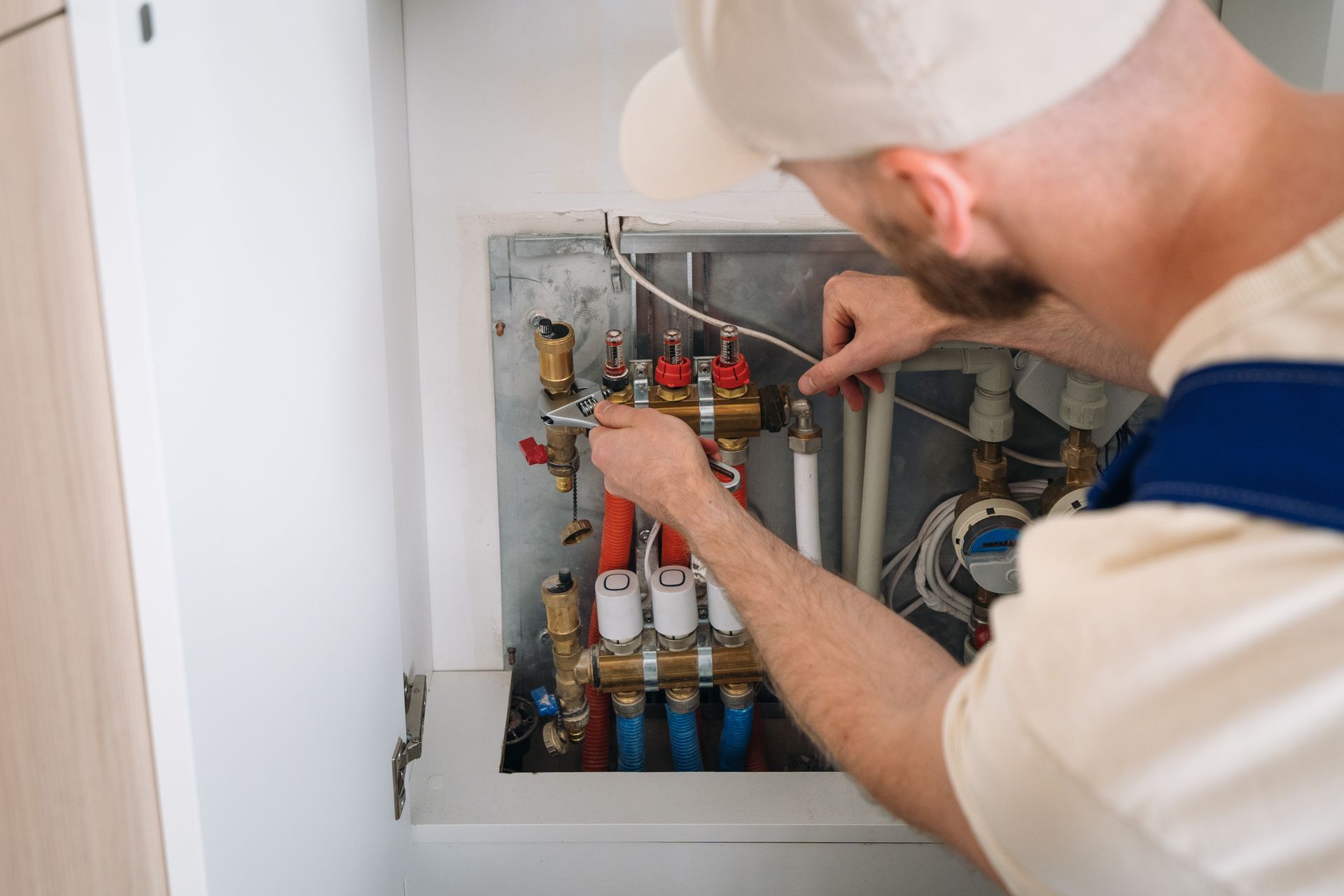 A plumber wearing a white cap and work clothes works on water pipes inside a cabinet.