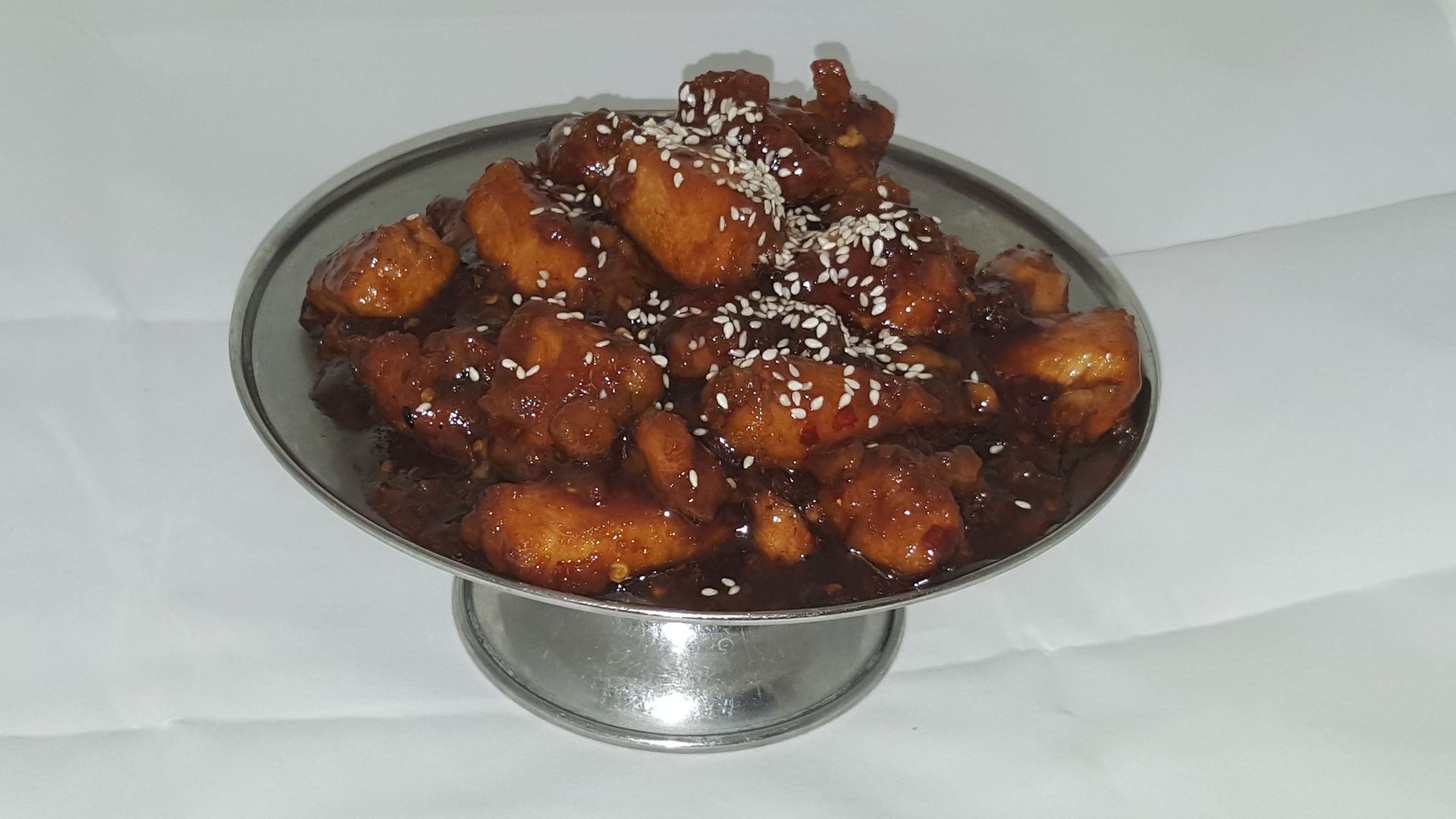 Sweet and sour chicken with sesame seeds in a metal serving dish on a white surface.