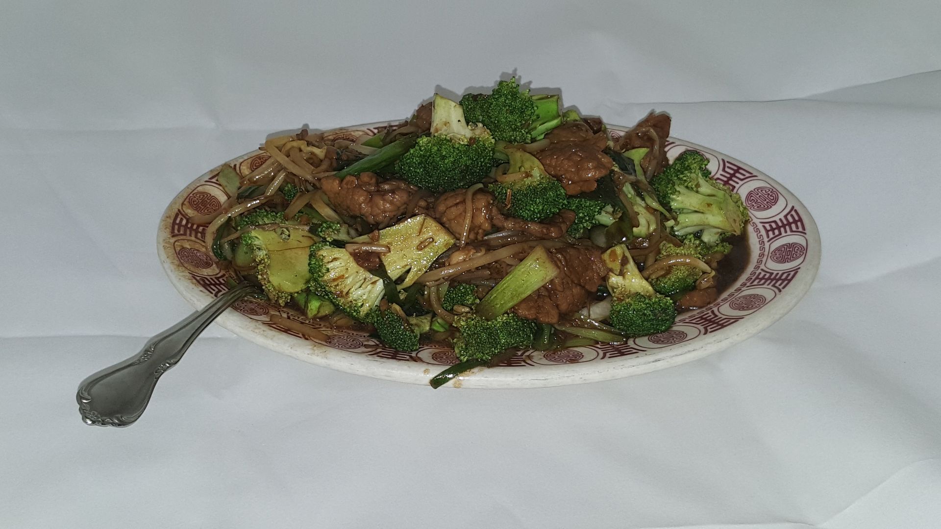 Beef and broccoli stir-fry on a patterned plate with a spoon, set on a white tablecloth.