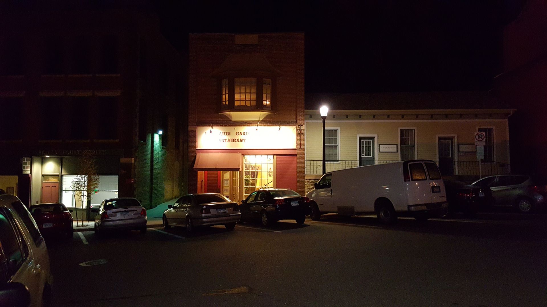 Night scene of buildings with lit storefronts, parked cars, and streetlights illuminating a dark street.