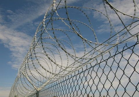 Chain-link fence topped with coils of razor wire against a blue sky with scattered clouds.