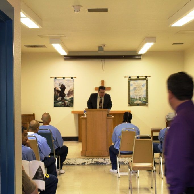 Prison chapel: A man in suit at a pulpit speaks to seated inmates, room lit by overhead lights and a crucifix.