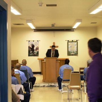 Prison chapel: A man in suit at a pulpit speaks to seated inmates, room lit by overhead lights and a crucifix.