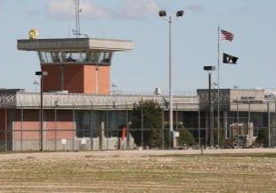 Prison with a watchtower, chain-link fence, and American flag on a sunny day.