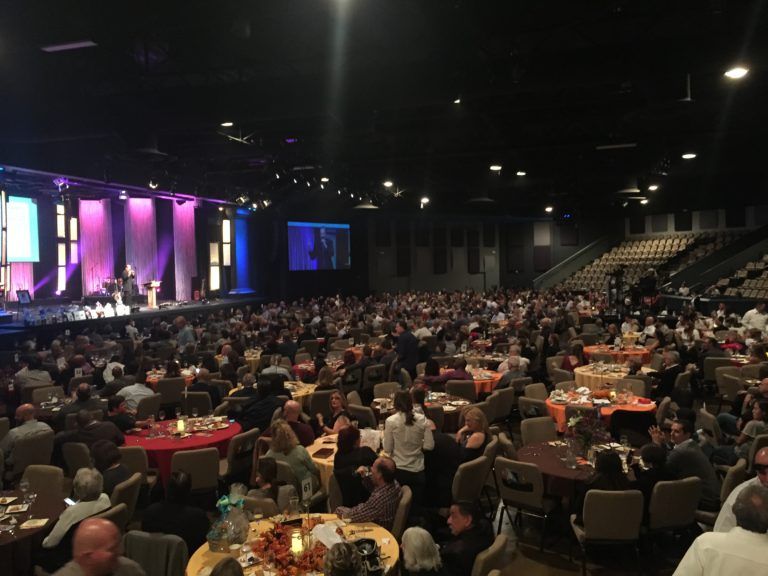 Large event hall with many people seated at round tables; stage at front with screens and performers.