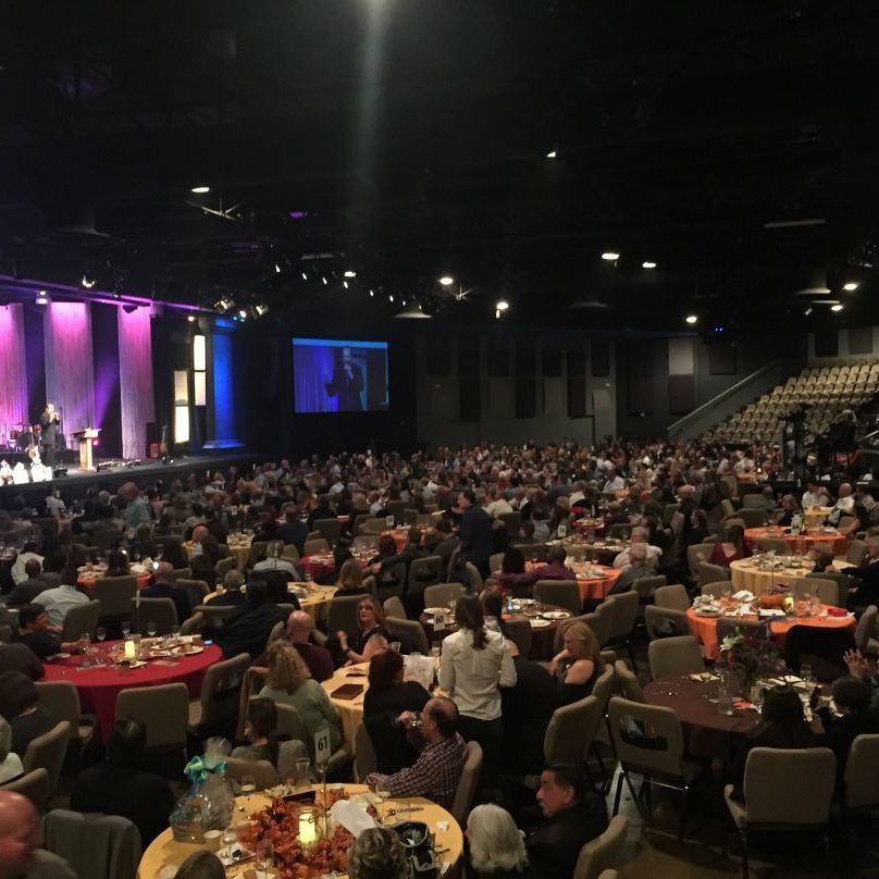 Large event hall with many tables filled with people; stage in front, screen, and elevated seating.
