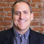 Man smiling, wearing a dark suit jacket over a blue button-down shirt, against a red brick wall.