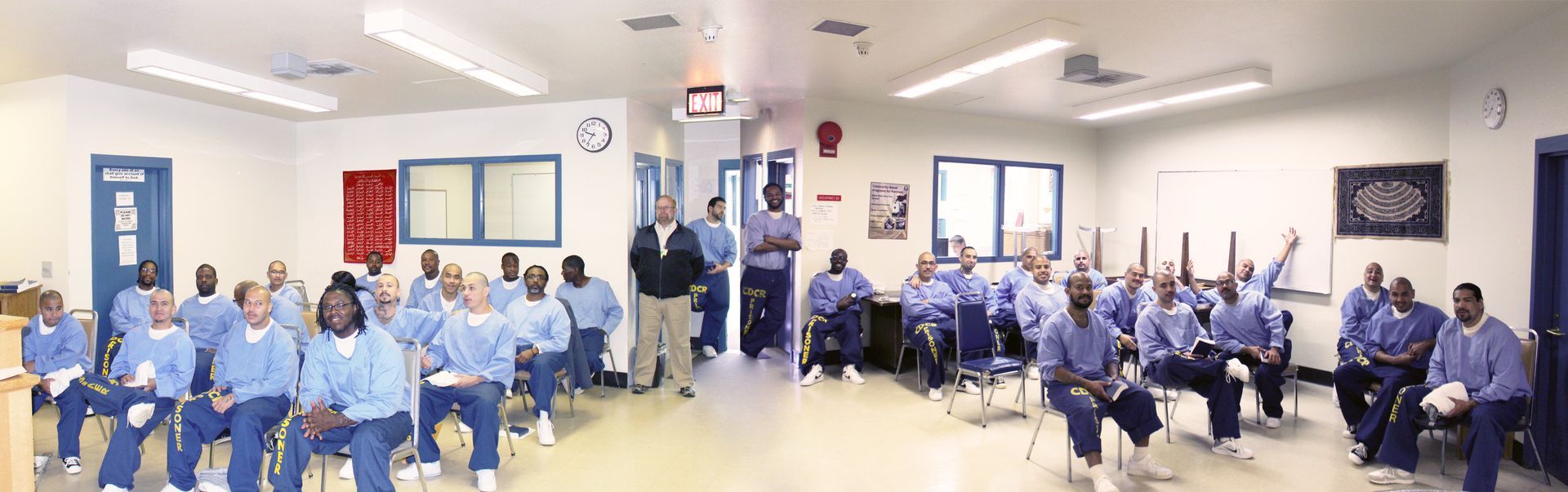 People in blue uniforms sit in chairs in a room. Some people stand at the front.
