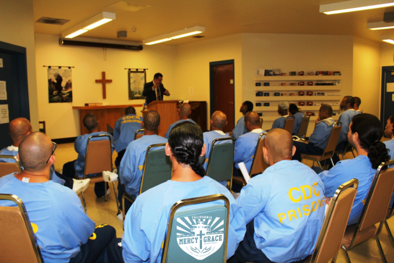 Men in blue uniforms attend a church service inside a room, with a pastor speaking at a podium.