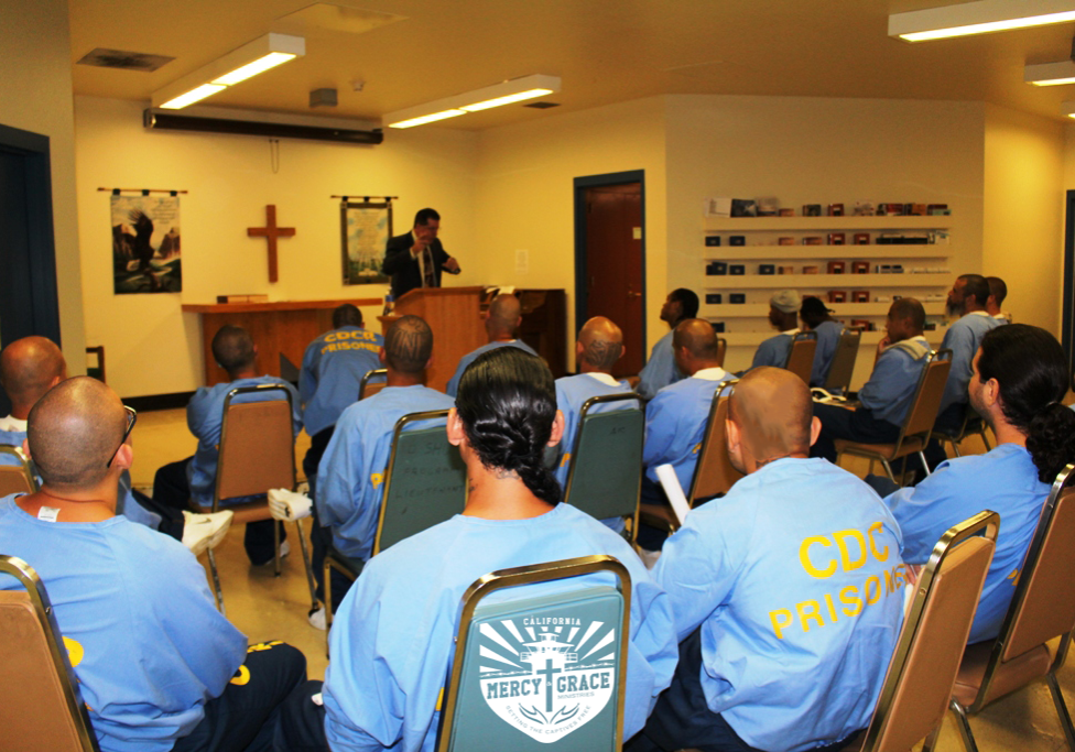 Men in blue prison uniforms attend a religious service in a room; a speaker stands at a lectern.