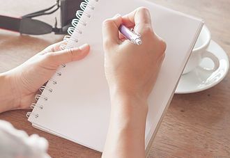 Person writing in spiral notebook with pen, sitting at a table with a camera and coffee cup nearby.