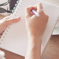 Hand holding a purple pen, writing in a white spiral notebook on a wooden surface.