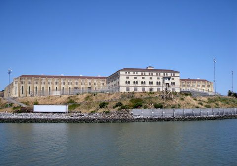 Stone building atop a hill near water; pale beige with brown roof, blue sky above.