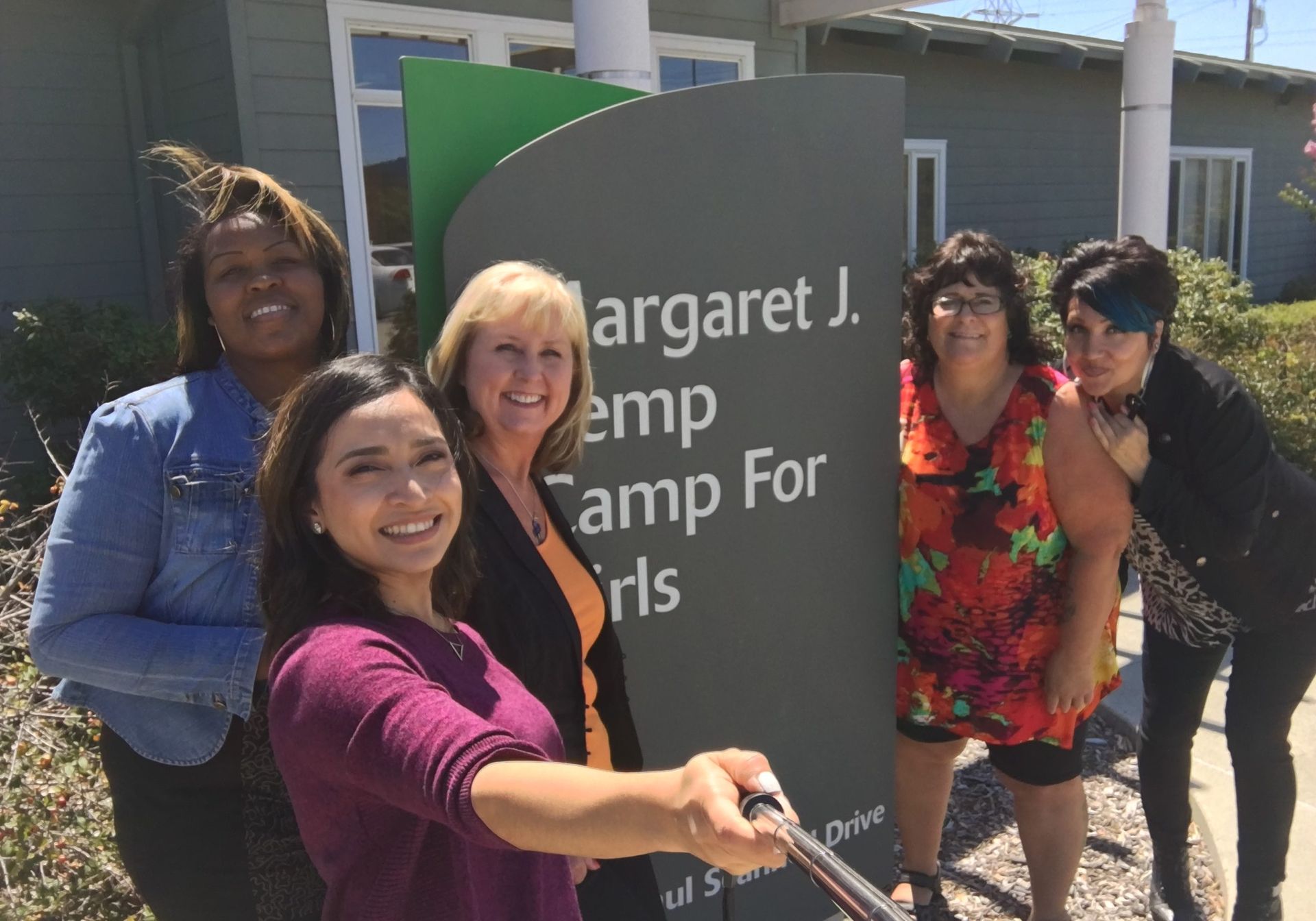 Five women pose with a sign for Margaret J. Kemp Camp For Girls. They are smiling outside on a sunny day.