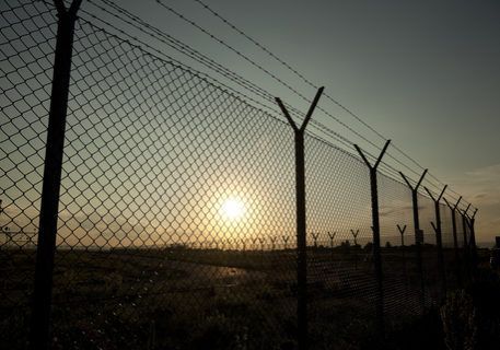 Chain-link fence with barbed wire silhouetted against a sunrise over a field.
