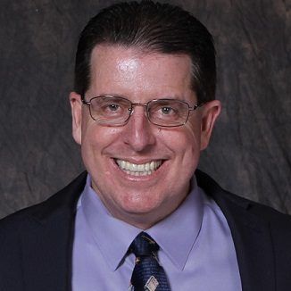 Man in glasses smiling, wearing a suit jacket and tie, against a dark gray backdrop.