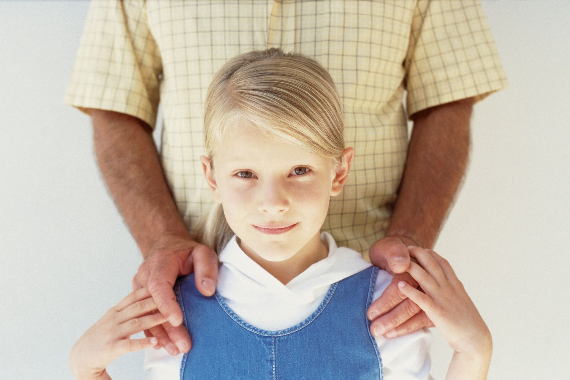 A person with their hands on the shoulders of a girl wearing a denim dress, white wall background.