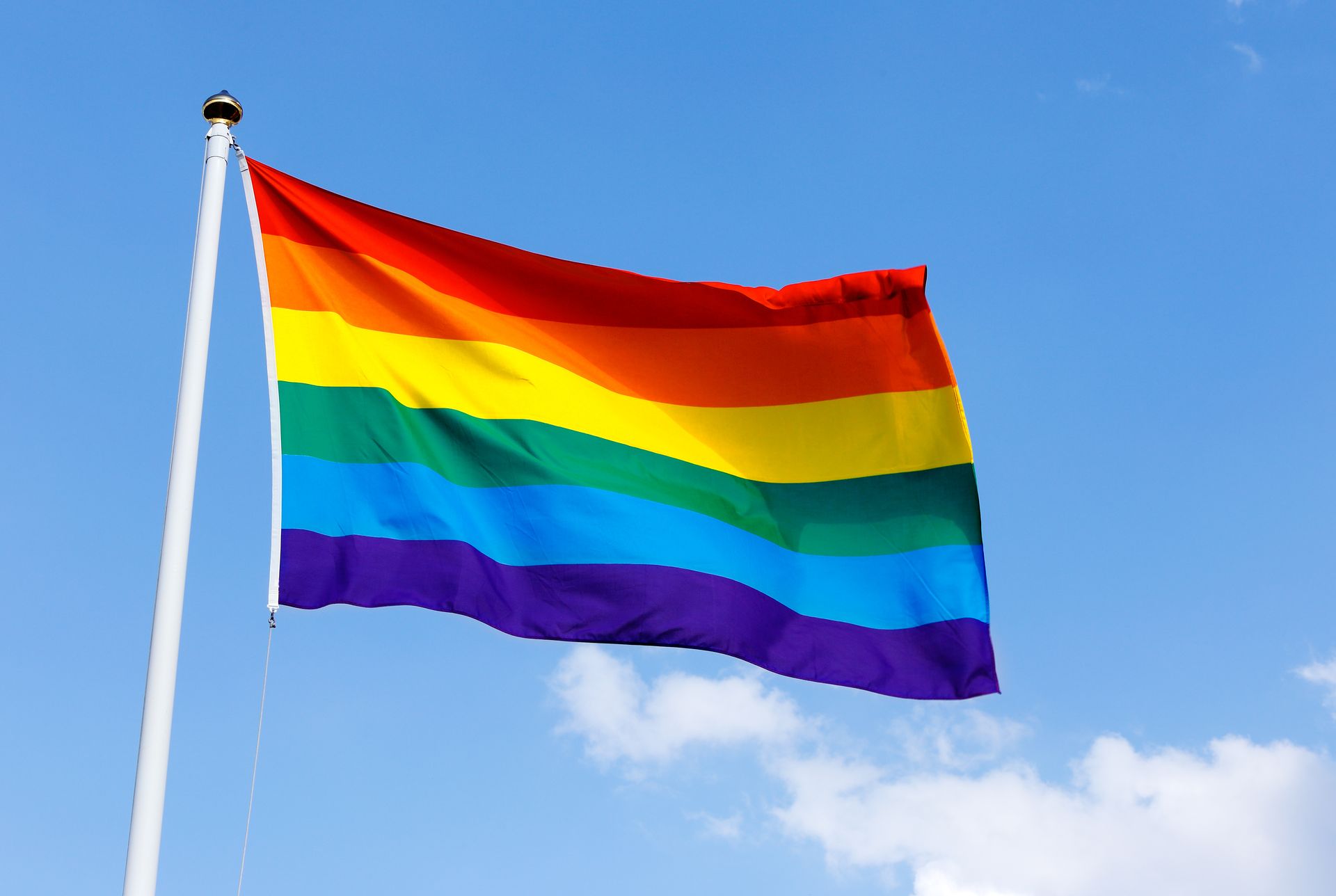 Rainbow Pride flag waving in a bright blue sky with wispy clouds.