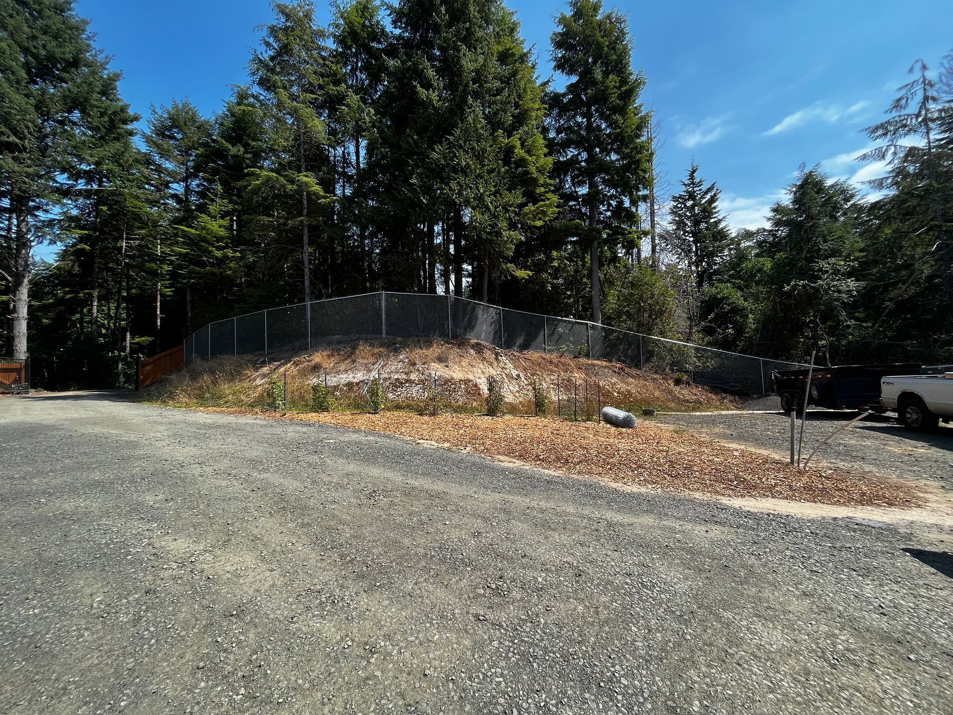 A white truck is parked in a gravel lot next to a fence surrounded by trees.
