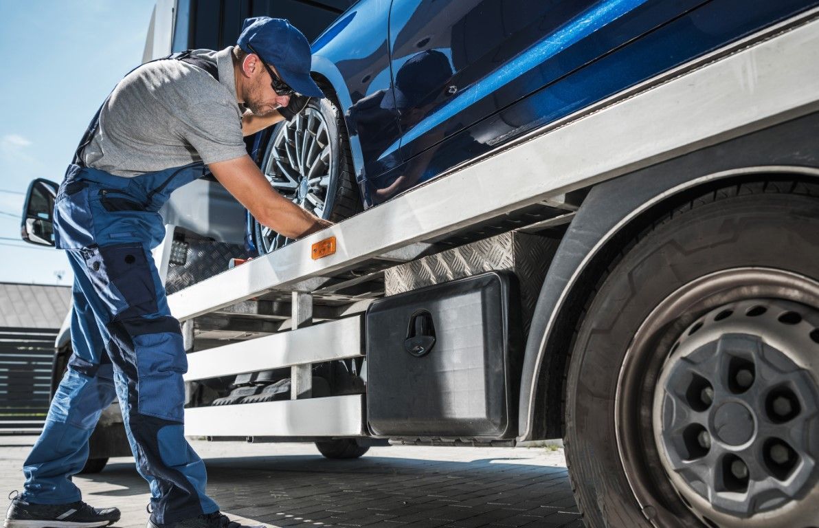 Tow truck operator securing a blue car to the flatbed. He is wearing overalls and a cap, working outdoors.