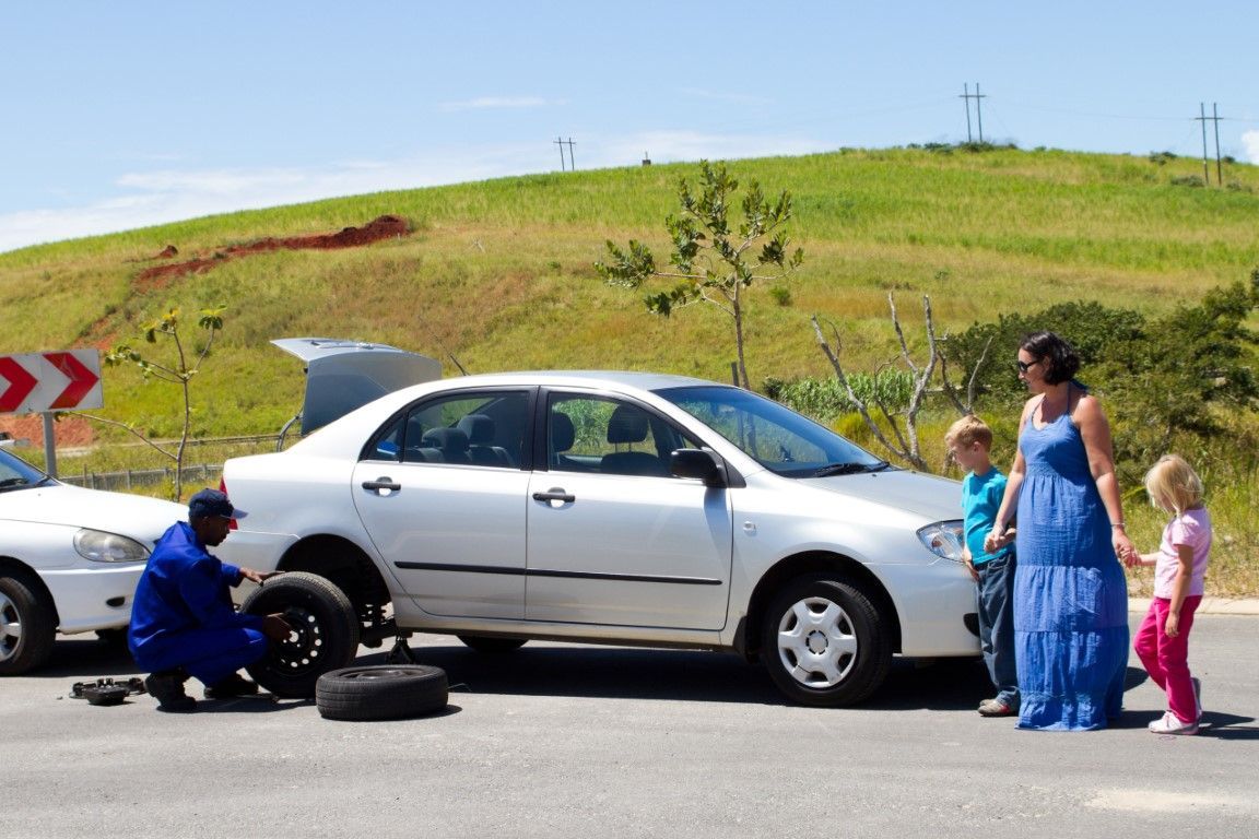 A car being repaired on the side of a road; a person in blue overalls is changing a tire; a woman and children watch.