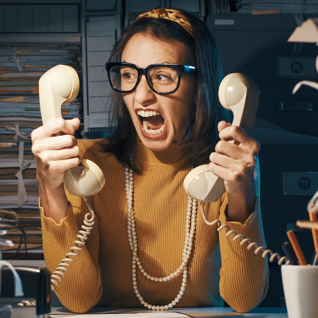 A woman wearing glasses is screaming while holding two telephones in her hands.