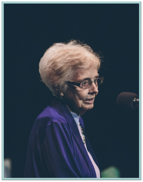 An elderly woman wearing glasses is speaking into a microphone