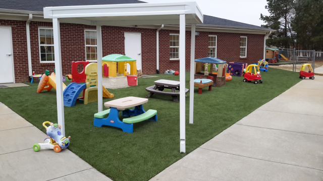 Playground with colorful toys and canopy, green turf, brick building in the background.