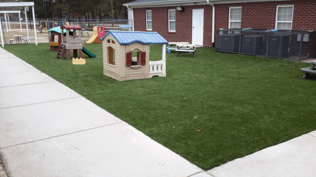 Playground with green artificial turf, playhouse, and brick building in background.