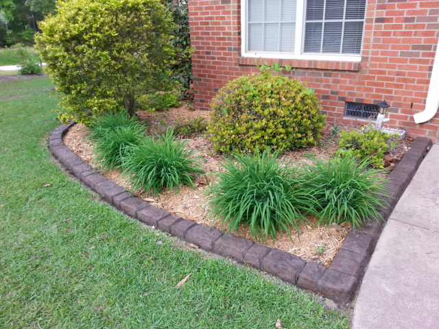 A corner flower bed with dark brown concrete curbing, green grass, and shrubs in front of a brick building.