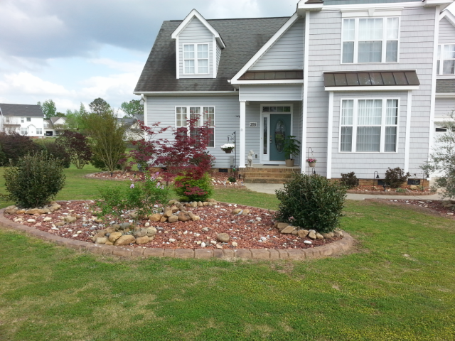 A light gray house with a red-rock landscaped front yard. Bushes and a small tree are in the yard.