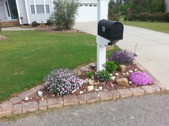 Black mailbox on white post with landscaped bed of flowers and rocks, in front of a house.