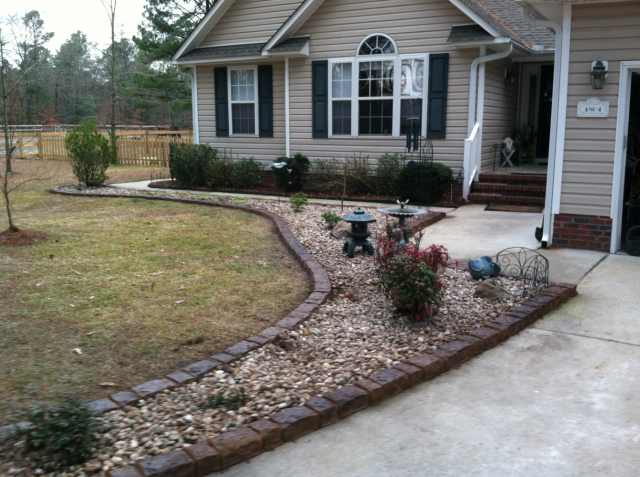 Tan house with a landscaped yard and stone-lined walkway filled with gravel and shrubs.