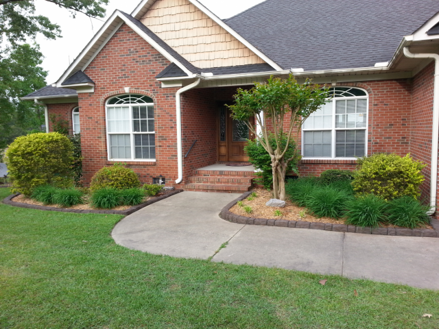 Red brick house with front yard; concrete path leads to the front door, flanked by greenery and bushes.